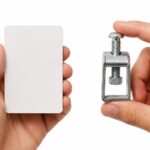 Duas mãos segurando lado a lado uma ficha de papel e um fixador metálico sobre fundo branco.